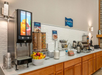 Breakfast buffet area with a juice dispenser, cereal containers, fruit bowl, waffle maker, coffee station, and condiments on a counter with wooden cabinets.