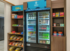 Hotel snack area with a fridge stocked with beverages, flanked by shelves holding snacks, chips, and medicine boxes.