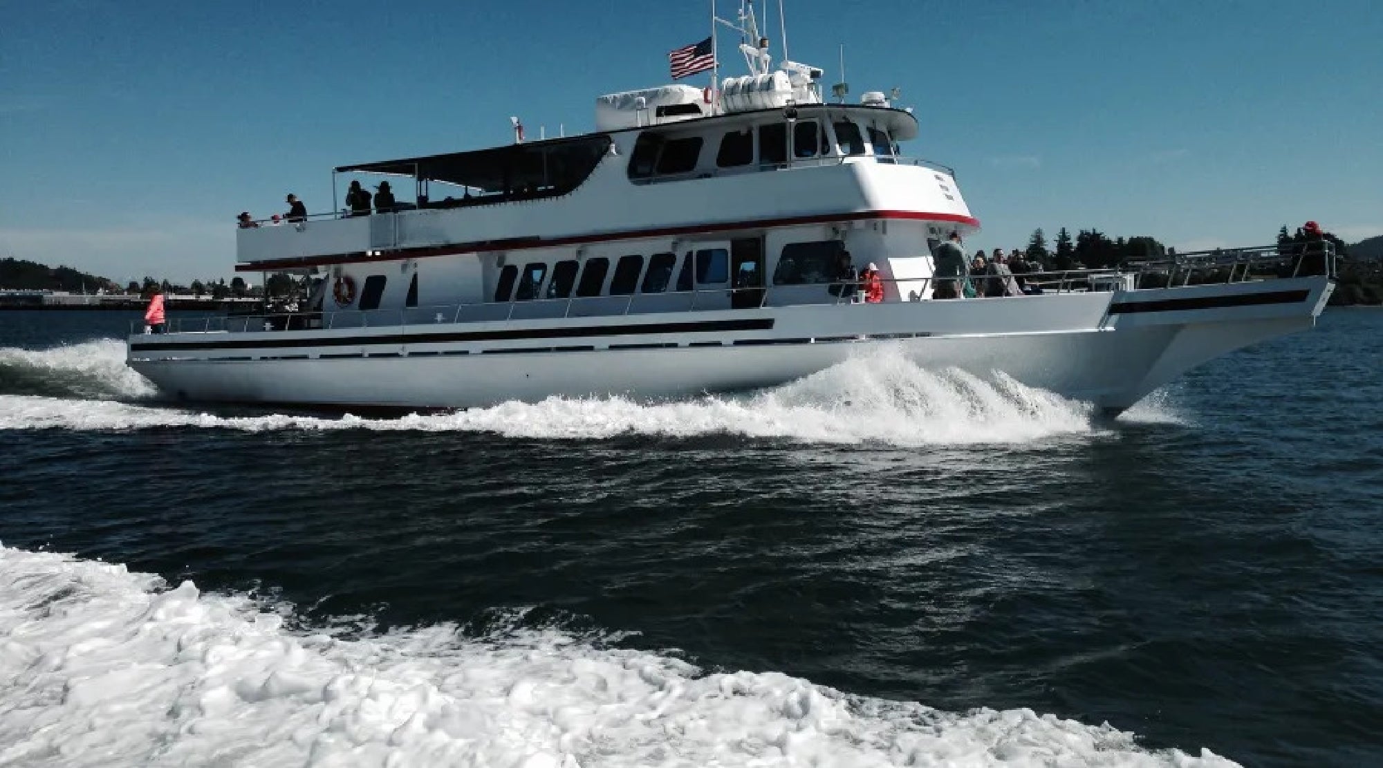 A large white ferry with a flag navigates through calm waters under a clear sky, with passengers visible on the upper deck.