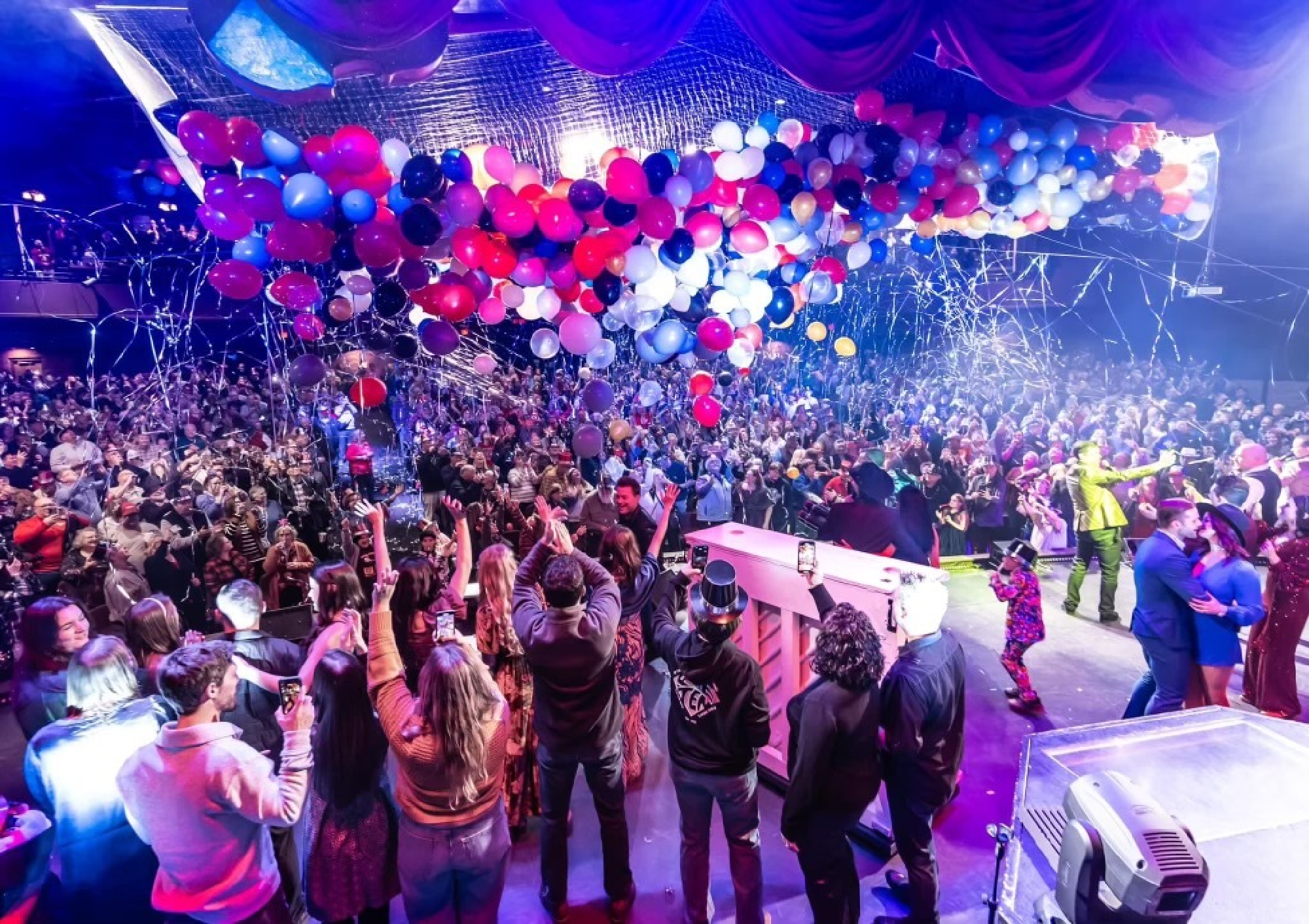 A lively crowd celebrates under colorful balloons and streamers in a festive indoor venue.