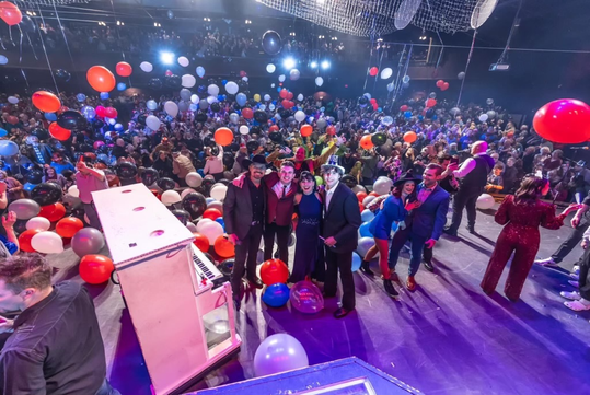 Large crowd at an indoor celebration with balloons, colorful lighting, and people dressed up.
