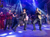 Group of people on stage celebrating New Year, with two men in the foreground dancing energetically. Stage lights and "Happy New Year" text in the background.