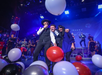 People celebrate on stage with balloons during a festive event. A man with a hat holds a microphone.