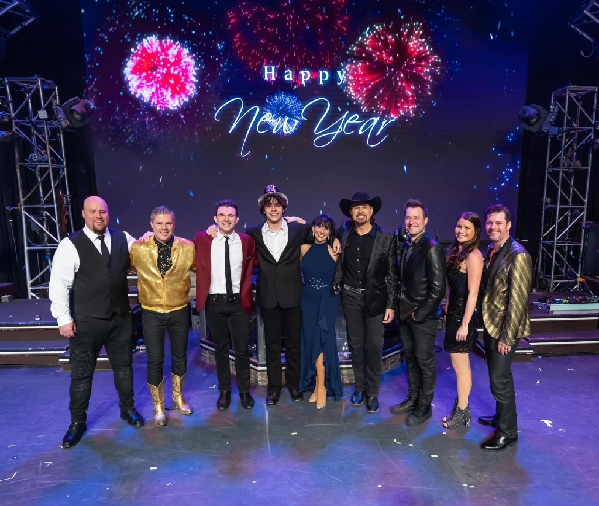 Nine people in festive attire stand on a stage under a "Happy New Year" sign, with fireworks displayed in the background.