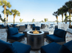 Outdoor patio with blue cushioned seating around a circular fire pit. Palm trees, tables, and umbrellas are visible. The background features a view of water and distant land.