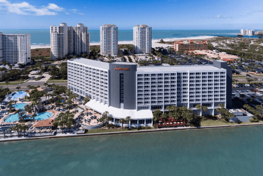 Aerial view of a large beachfront hotel with multiple floors, surrounded by water and nearby high-rise buildings. Several pools and a parking area are visible.