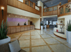 Hotel lobby with a reception desk, two employees, and a seating area. The room features tiled floors, potted plants, and a staircase leading to an upper level.