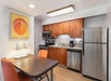 A compact kitchen with wooden cabinets, stainless steel appliances, a table with two chairs, and a grapefruit artwork on the wall.