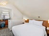 A neatly made bed with white linens sits against a slanted wall; an open doorway reveals a bathroom vanity with a mirror and lights.