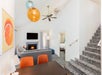 Modern hotel suite with loft. Features a living area with sofa, TV, fireplace, and artwork, and a dining table with orange chairs. Carpeted stairs lead to an upper level. Bedroom visible in back.