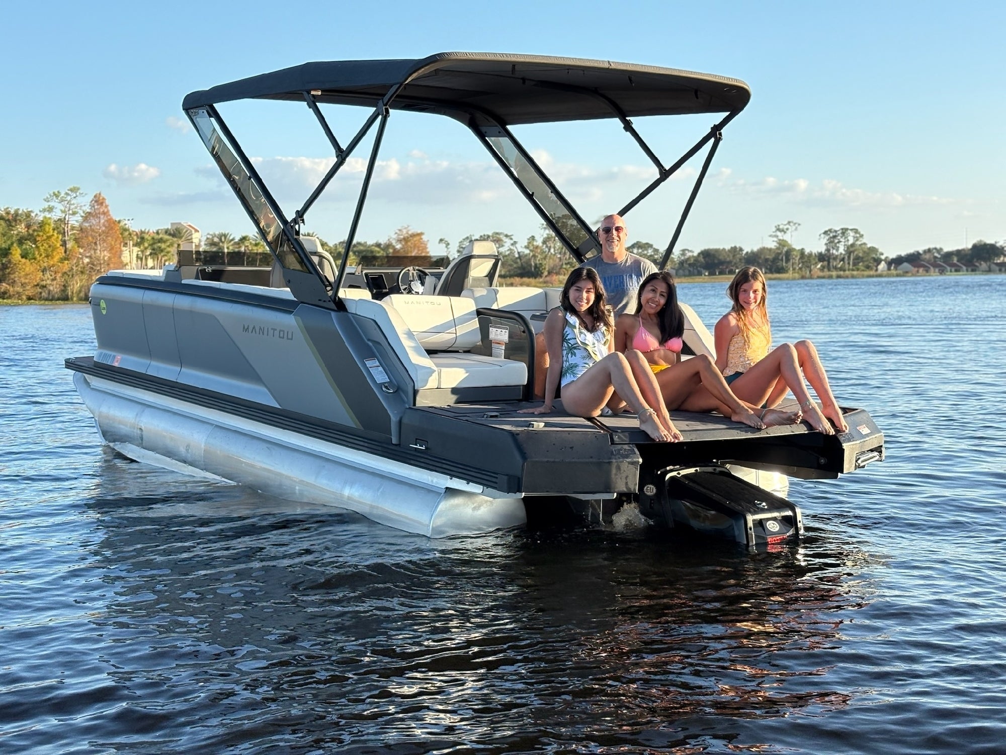 Three women sit with their feet in the water at the back of a pontoon boat, while a man stands near the helm. The boat is on a calm lake under a clear sky.