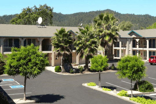 Exterior of the Cloverdale Wine Country Inn & Suites on a sunny day with several trees in the parking lot and in front of the building.