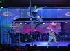 Performers on a stage, one balancing on a vertical pole and another climbing it, with an audience watching. A person in the foreground holds a microphone near a booth labeled "Telephone".