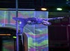 A performer wearing a shiny black shirt and jeans hangs upside down on a pole against a colorful, patterned background during a stage performance.