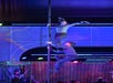 Two performers in casual clothing are balancing on a pole on stage, with colorful lights and a telephone booth in the background.