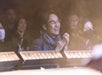 Person clapping and smiling in an audience, surrounded by other people, some wearing masks.