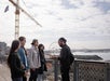 A group of four people listens to a man speaking outdoors near a waterfront, with a ferris wheel and construction crane in the background.
