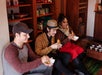 Three people sit indoors near shelves, smiling and eating snacks from bowls. Cups and teapots are arranged on the floor in front of them.