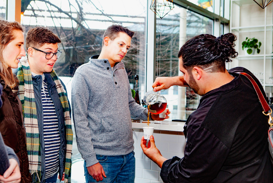 A man pours tea into a cup held by another man while two people watch in a bright, modern cafe setting.
