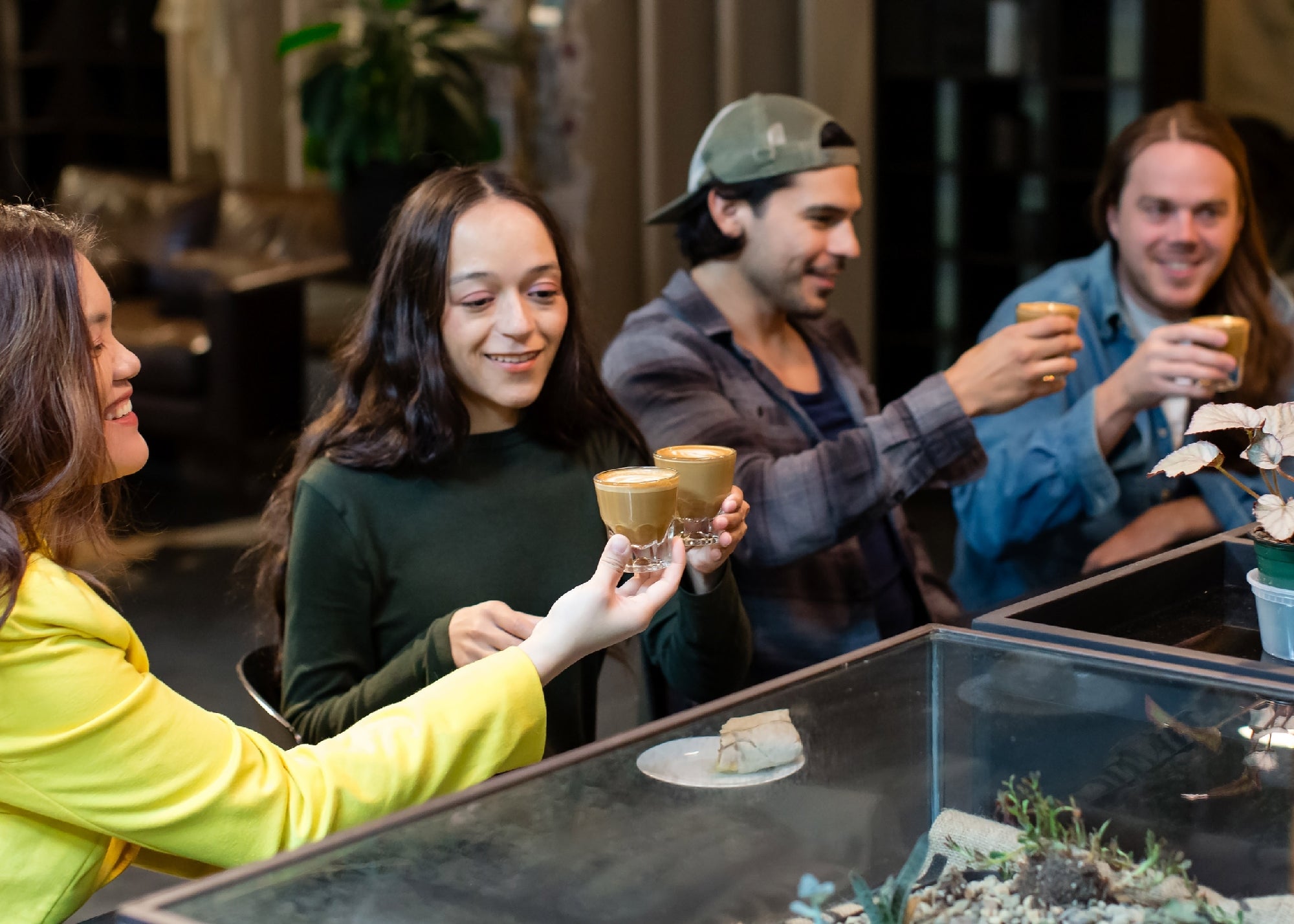 Four people sitting at a table, smiling and holding up coffee glasses in a toast gesture in a modern indoor setting.