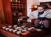 A person wearing a shawl pours tea from a black teapot into small cups arranged on a wooden table in a cozy indoor setting.