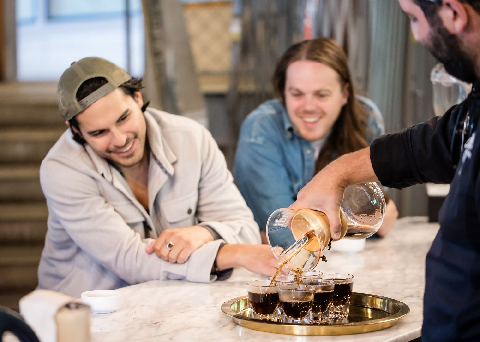 A barista pours coffee from a glass carafe into cups on a tray while two men sitting at the counter watch and smile.
