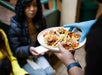 A person wearing blue gloves serves a plate of tacos to a seated woman holding a white napkin.