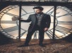 Man in a dark suit holds a deerstalker hat, standing in front of a large clock face with Roman numerals.