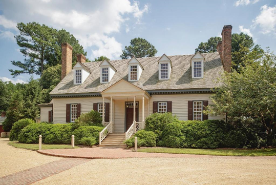 A two-story colonial-style house with a gabled roof, dormer windows, and a central entrance. Surrounded by trees and bushes, it has a brick walkway leading to the front steps.