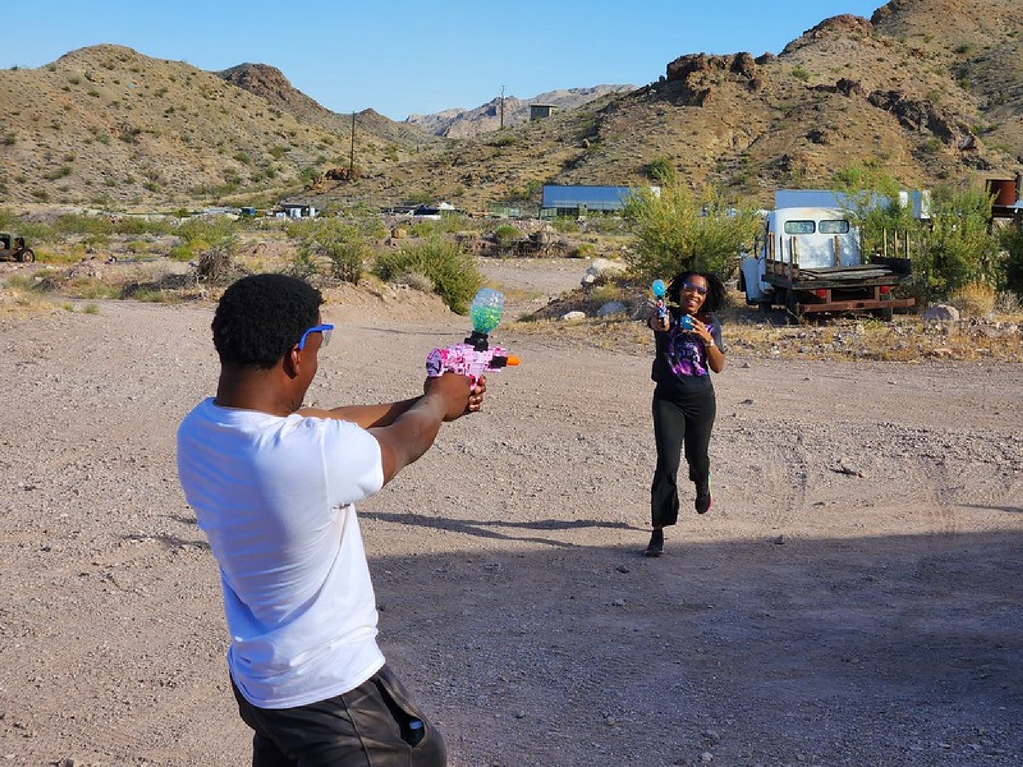 Two people playing with water guns in a rocky, desert landscape. The person in the foreground aims at the other, who is running and smiling.