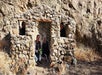 Four people sit and stand inside a small stone structure with no roof, set against a rocky hillside. Dry grass surrounds the area.