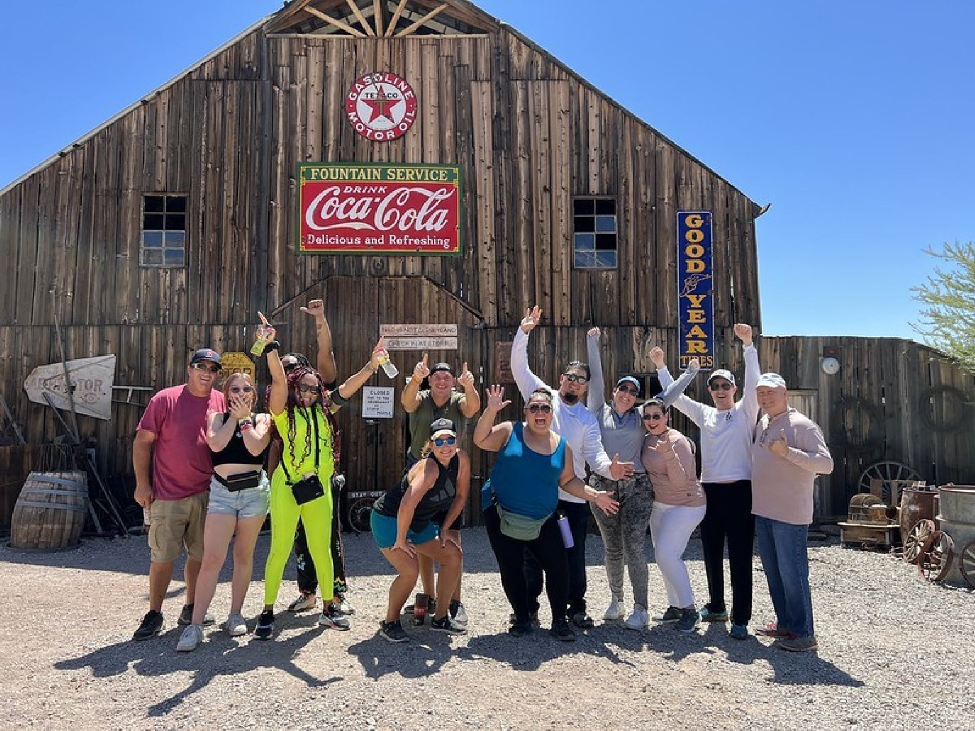 A group of people pose cheerfully in front of a rustic wooden building with vintage Coca-Cola and Goodyear signs.