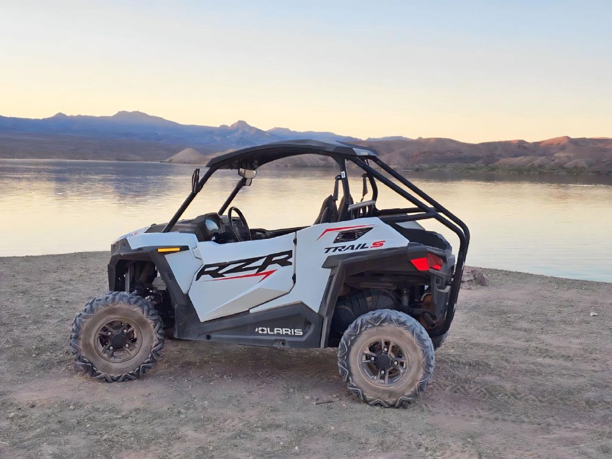 A white Polaris RZR off-road vehicle is parked on a dirt path near a calm lake with distant mountains under a clear sky.