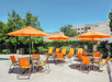 Outdoor patio with multiple orange tables and chairs under matching umbrellas, surrounded by greenery and a building in the background.