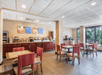 Hotel breakfast area with empty tables and chairs; self-serve counter includes coffee machines, cereal dispensers, and juice station with wall art above.