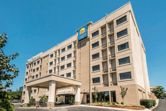 Six-story beige hotel building with blue and yellow signage, surrounded by small trees and landscaped greenery, under a clear blue sky.