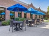 Outdoor patio with brown tables and chairs under blue umbrellas, set near a brick building and green shrubs.