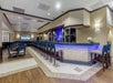 A modern bar and dining area with blue-lit counter, high stools, tables, and chairs. Wood and tile flooring, neutral-toned walls, and a large TV screen adorn the space.