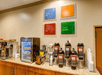 A coffee station with several different types of coffee in dispensers with cups to the right and a mini fridge with other drink options to the left.