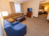 Hotel room with a seating area featuring a brown sectional sofa, blue chair, TV, and a view into a bedroom with a large bed and lamp.