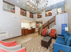 Hotel lobby with a chandelier, reception desk, and seating area. Features a large patterned rug, blue and red chairs, and a brown ottoman. Stairs lead to an upper level with a balcony.