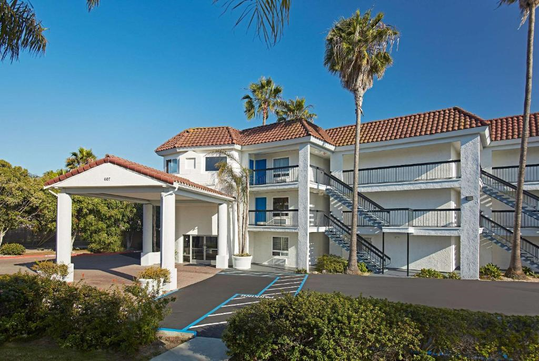 A white, three-story building with a red-tiled roof, palm trees, and an entrance canopy. Exterior staircases are visible alongside the structure, with a parking area in front.
