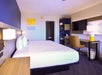 Hotel room with a large bed, two bedside lamps, a wall-mounted TV, desk with chair, a small fridge, and a colorful painting on the wall.