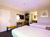 Hotel room with two double beds, a desk and chair, wall-mounted TV, and a small kitchenette area. Art piece and lamp on the wall.