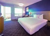 A hotel room with a large bed, blue accent wall, bedside tables with lamps, a wall painting, and an open door leading to a balcony with outdoor light.