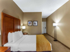 Hotel room with a neatly made double bed, wooden headboard, two framed artworks on the wall, a bedside lamp, and a floor lamp near the entrance door.