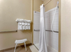 Shower with a white curtain, grab bars, and a handheld showerhead. Towels are stacked on a wall rack, and a small bench is placed nearby.