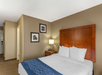 Hotel room with a neatly made double bed, wooden headboard, nightstand with a lamp, and framed pictures on the beige walls.