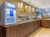 Breakfast buffet area with a refrigerator, serving trays with pastries, chafing dishes, cereal dispensers, and beverage station on a wooden counter in an airy room.