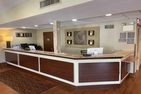 Hotel reception desk with brown wood paneling, computers on the counter, modern wall art, and a brown carpeted floor.